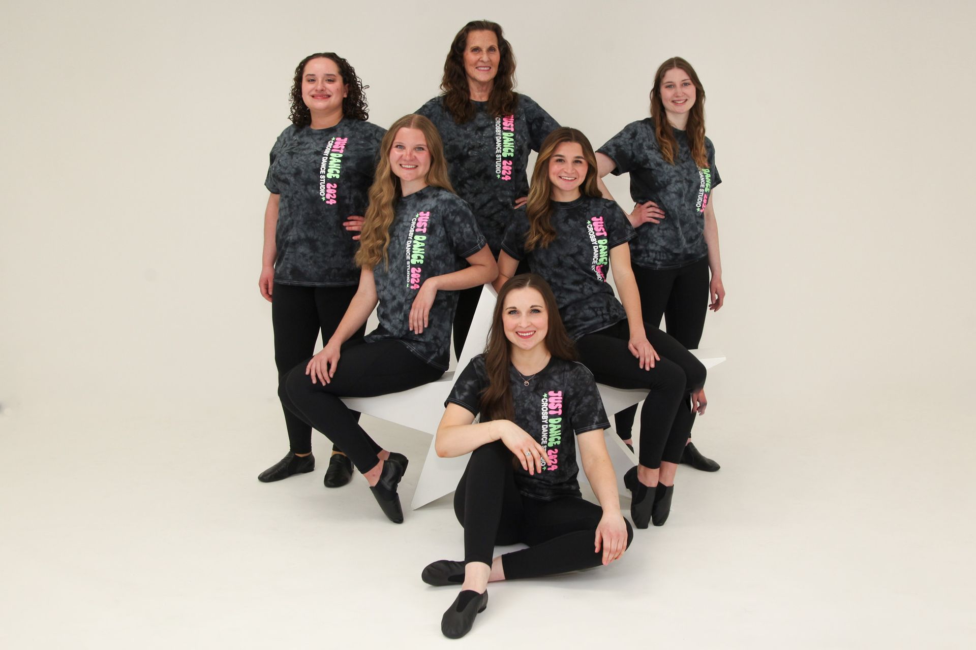 Seven people in matching dark shirts pose on white steps against a white backdrop.