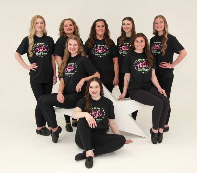 Seven dancers in matching black outfits pose on white steps in a studio setting.