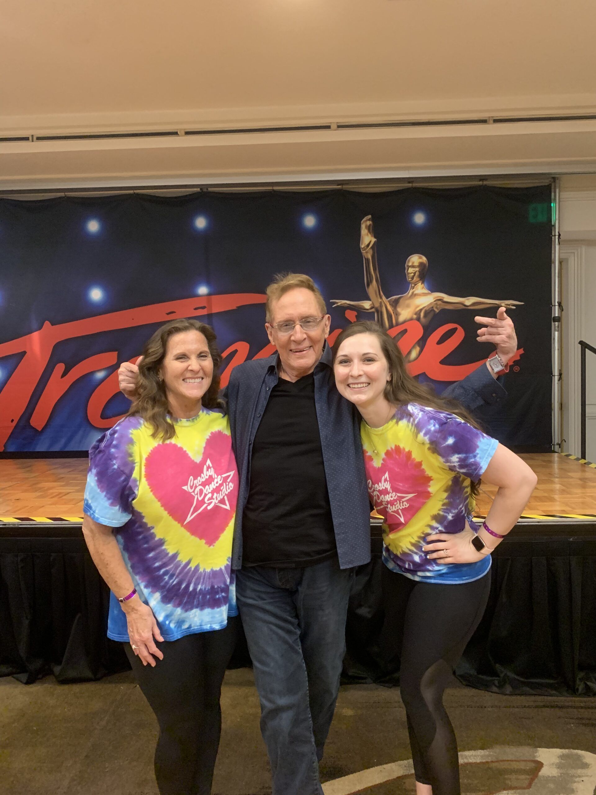 Three smiling people in tie-dye shirts posing on a stage with a black backdrop and red lettering