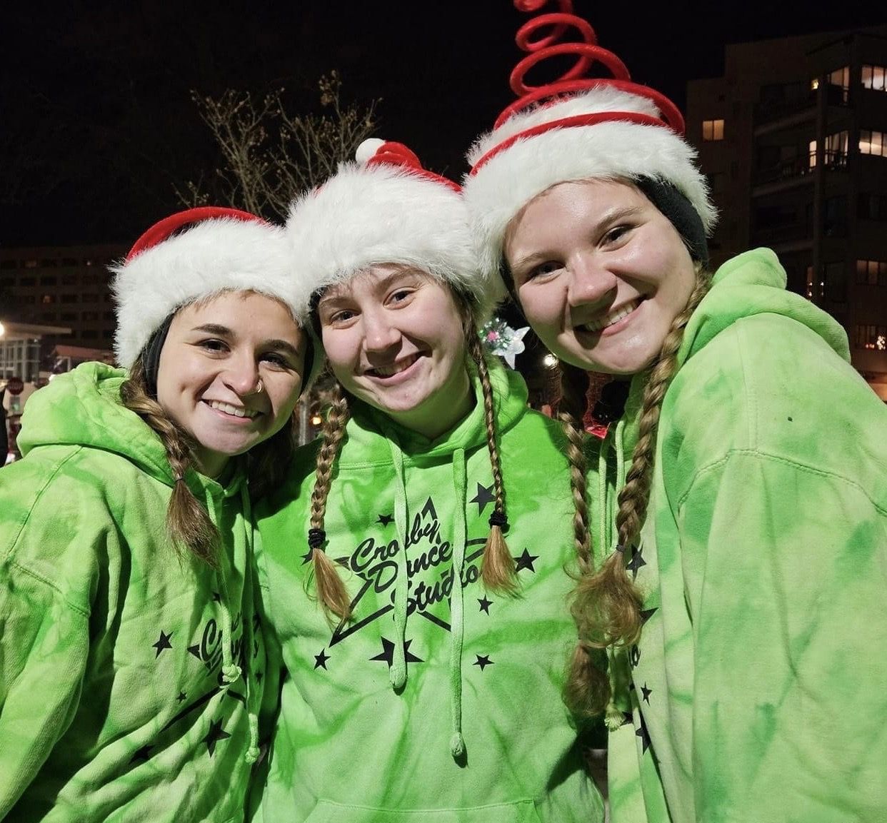 Three people in green hoodies and Santa hats smiling outdoors at night.
