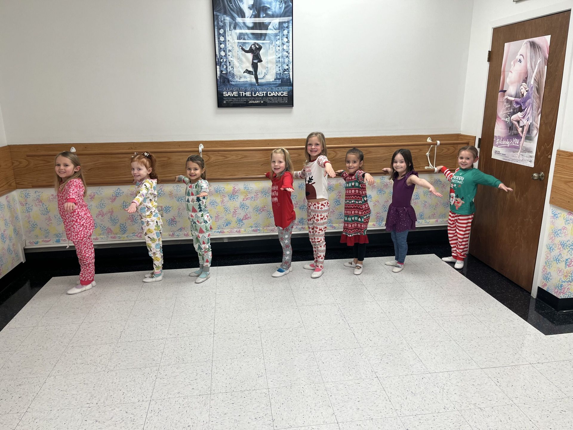 Seven children standing in a line against a hallway wall, arms out, in colorful pajamas.