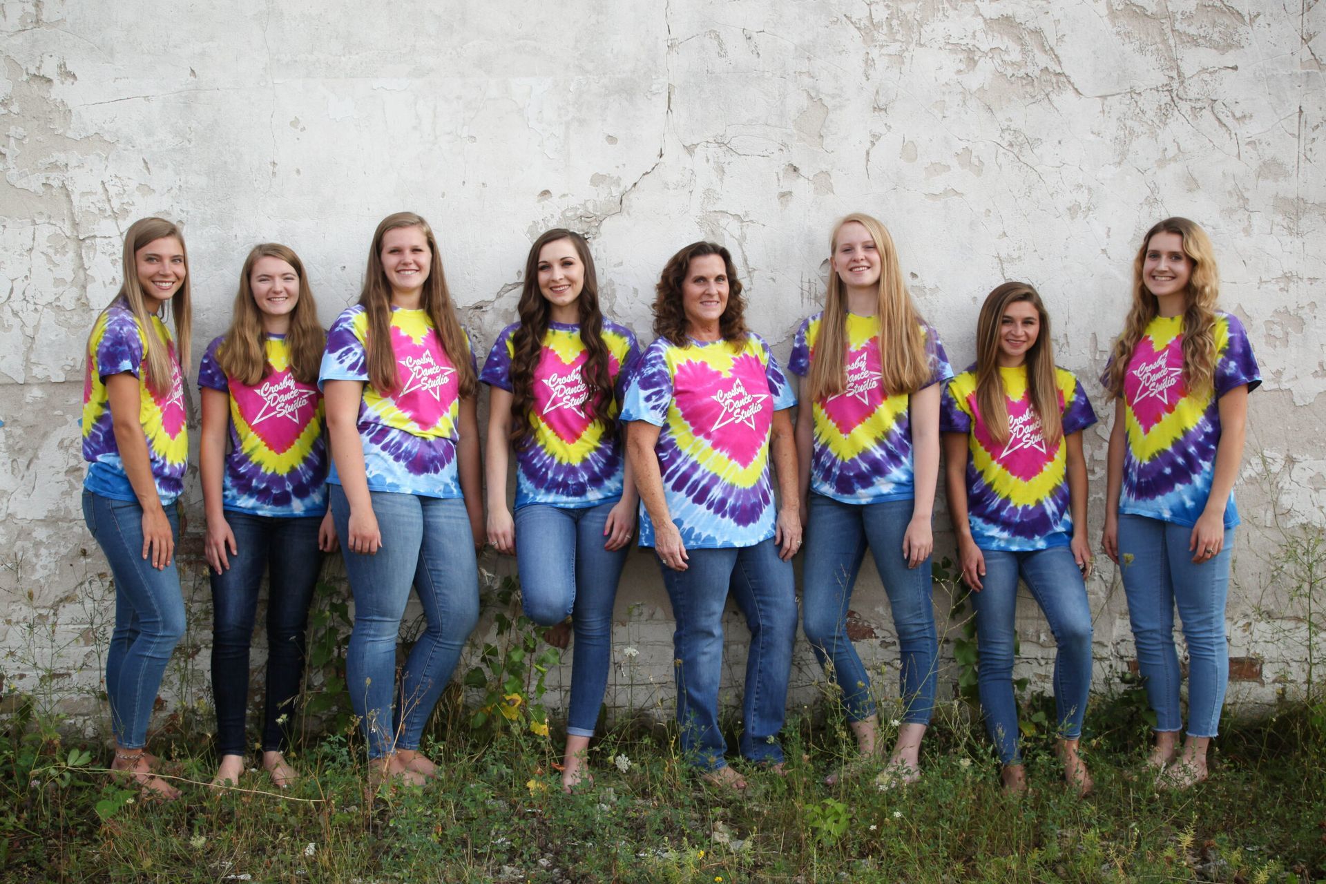 Eight people in matching colorful shirts and jeans standing against a weathered wall