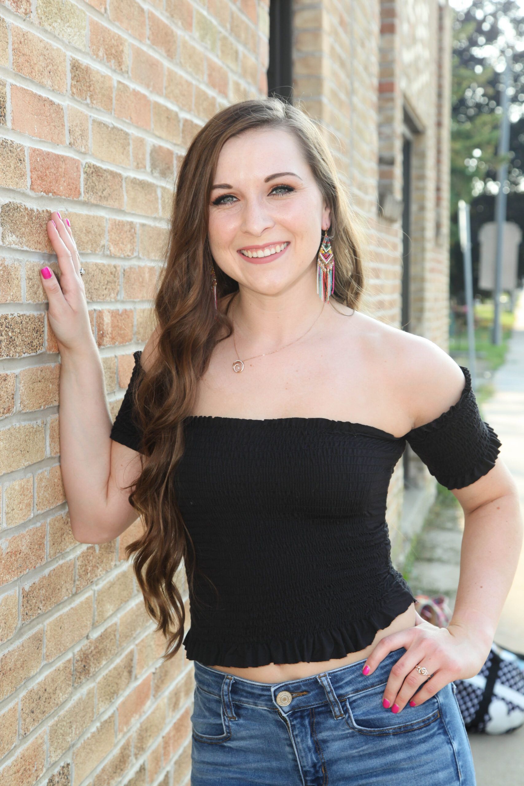 Smiling woman in a black off-shoulder top and jeans leaning against a brick wall outdoors