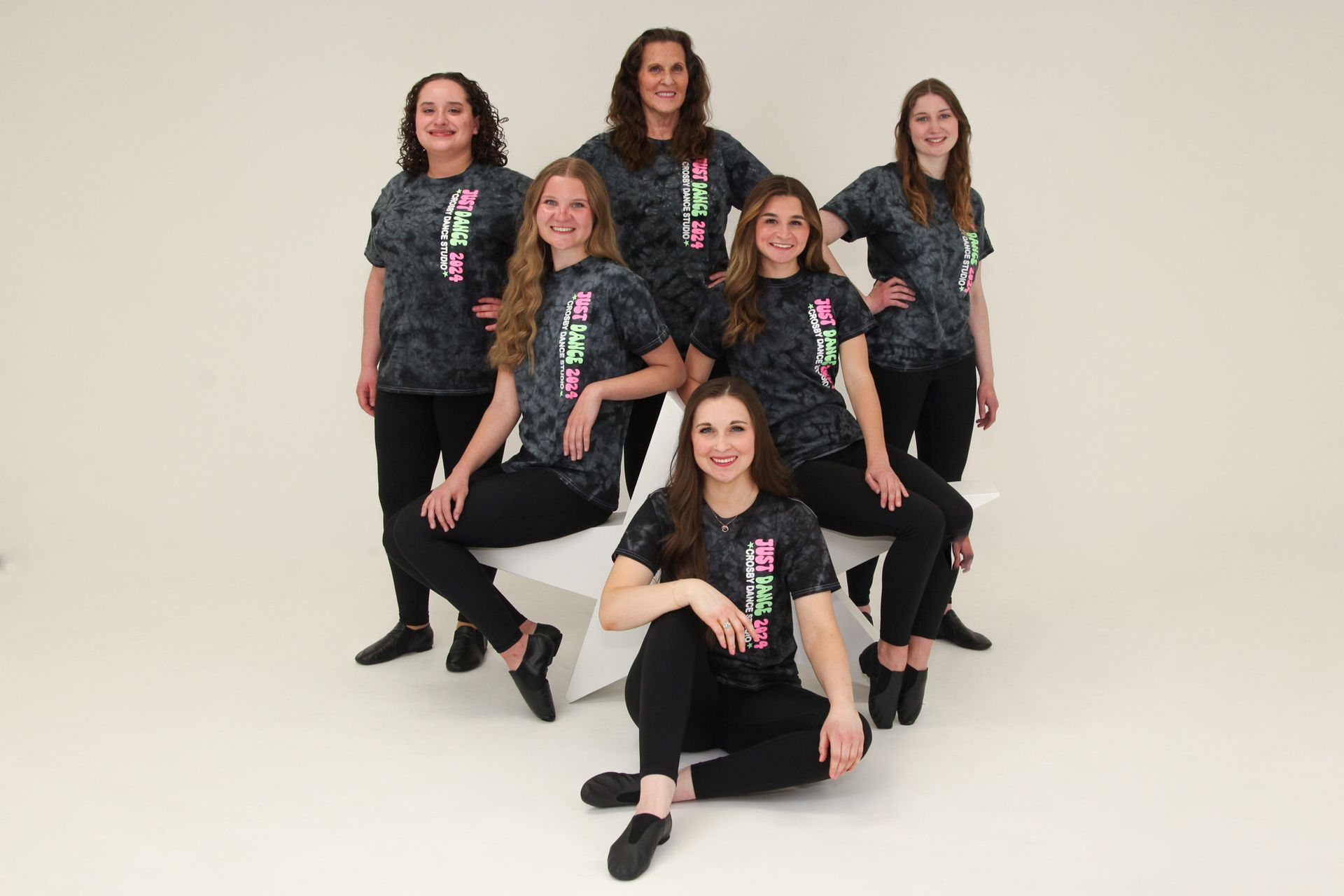 Seven dancers in matching black outfits pose on white steps in a studio setting.