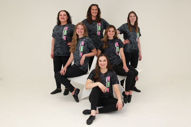 Seven dancers in matching black outfits pose on white steps in a studio setting.