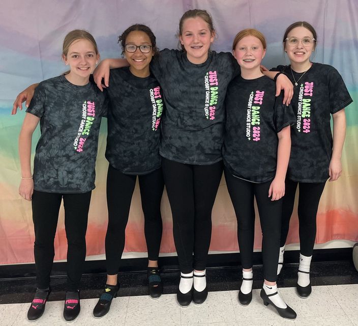 Five girls in matching dark shirts and black leggings stand arm-in-arm against a colorful wall.