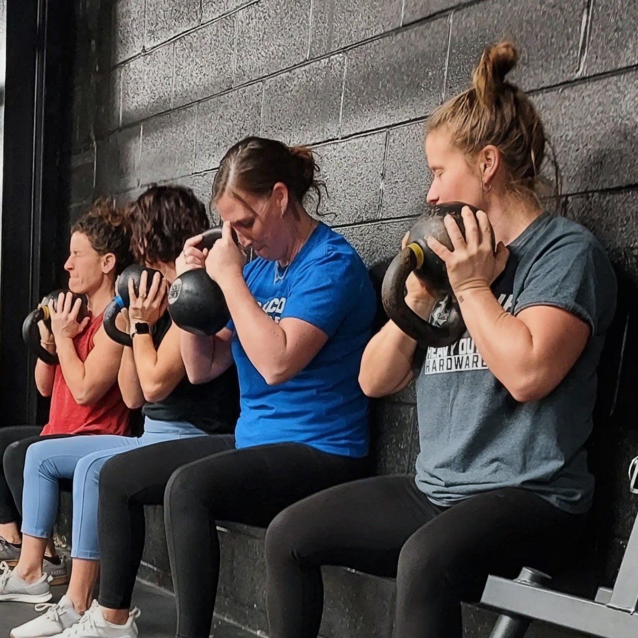 People in a fitness class at Vitality Athletics in Ephrata, PA hold kettlebells, seated against a black wall.