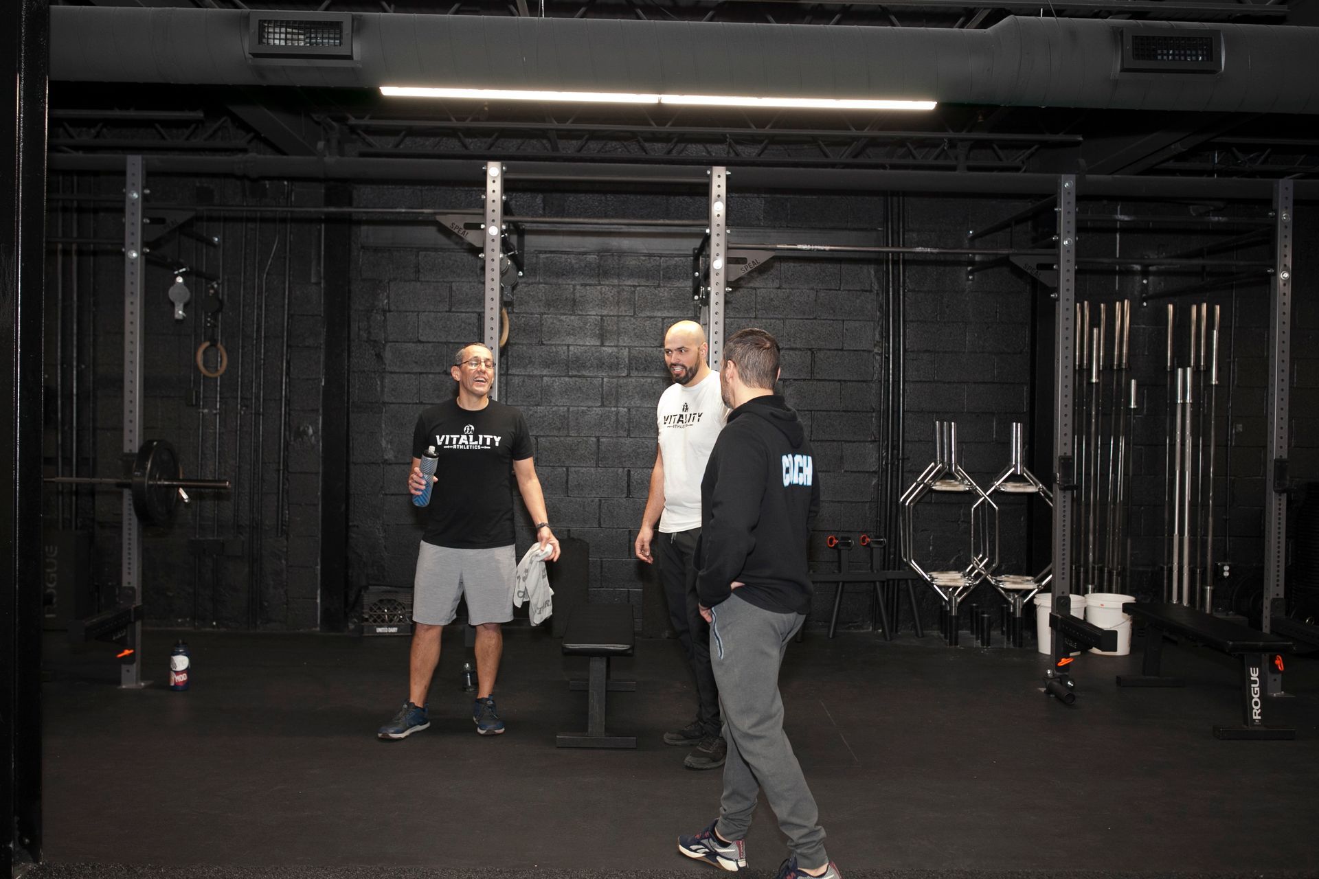 Three men in Vitality Athletics in Ephrata, PA, two talking, one holding a towel. Black bars, dark setting.