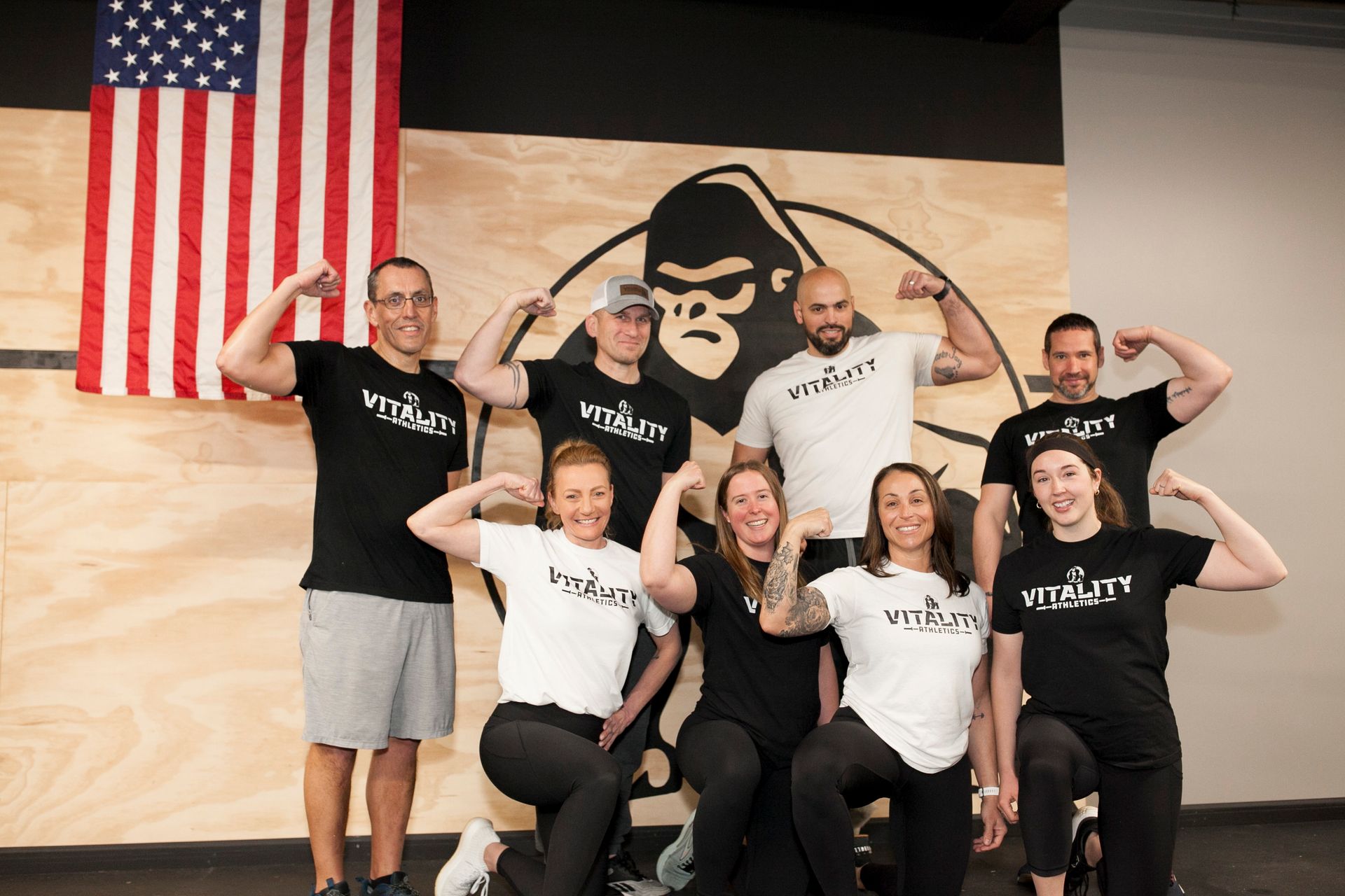 Group flexing in front of a gorilla logo and American flag at Vitality Athletics in Ephrata, PA. Gym setting, black and white shirts, smiling.