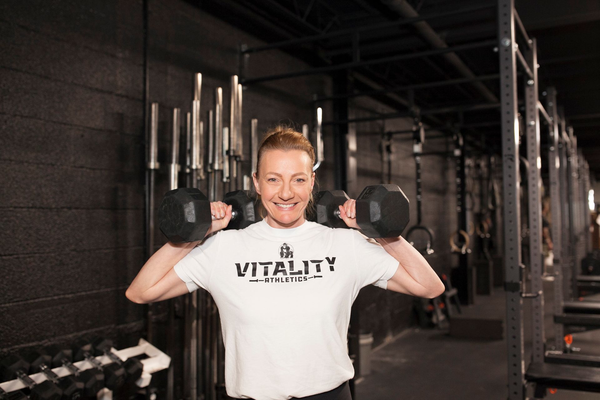 Woman in white t-shirt lifting dumbbells in Vitality Athletics in Ephrata, PA. Smiling, black wall background.