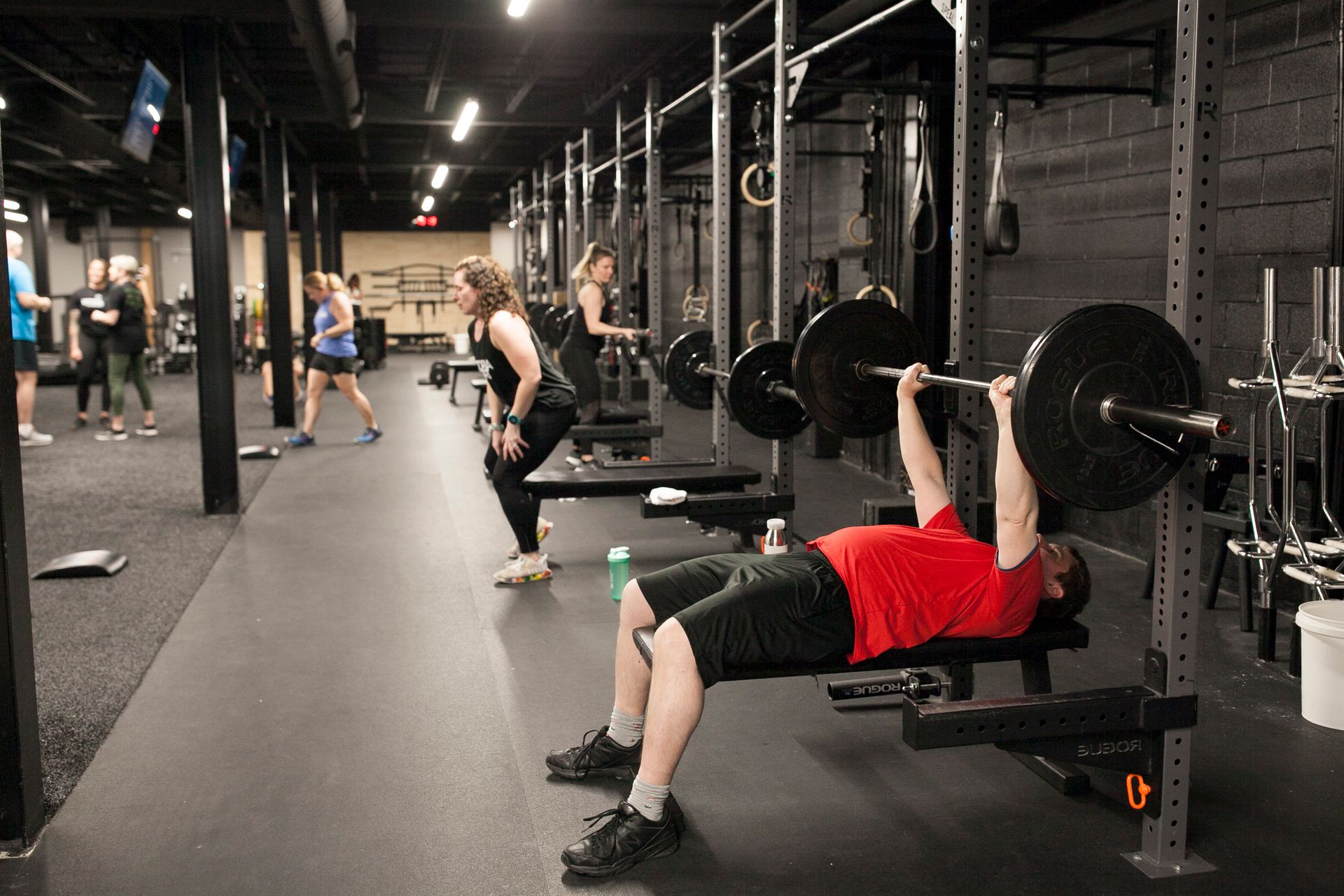 Man bench presses weight bar in Vitality Athletics in Ephrata, PA. Other people exercise in the background. Black gym with dark flooring.