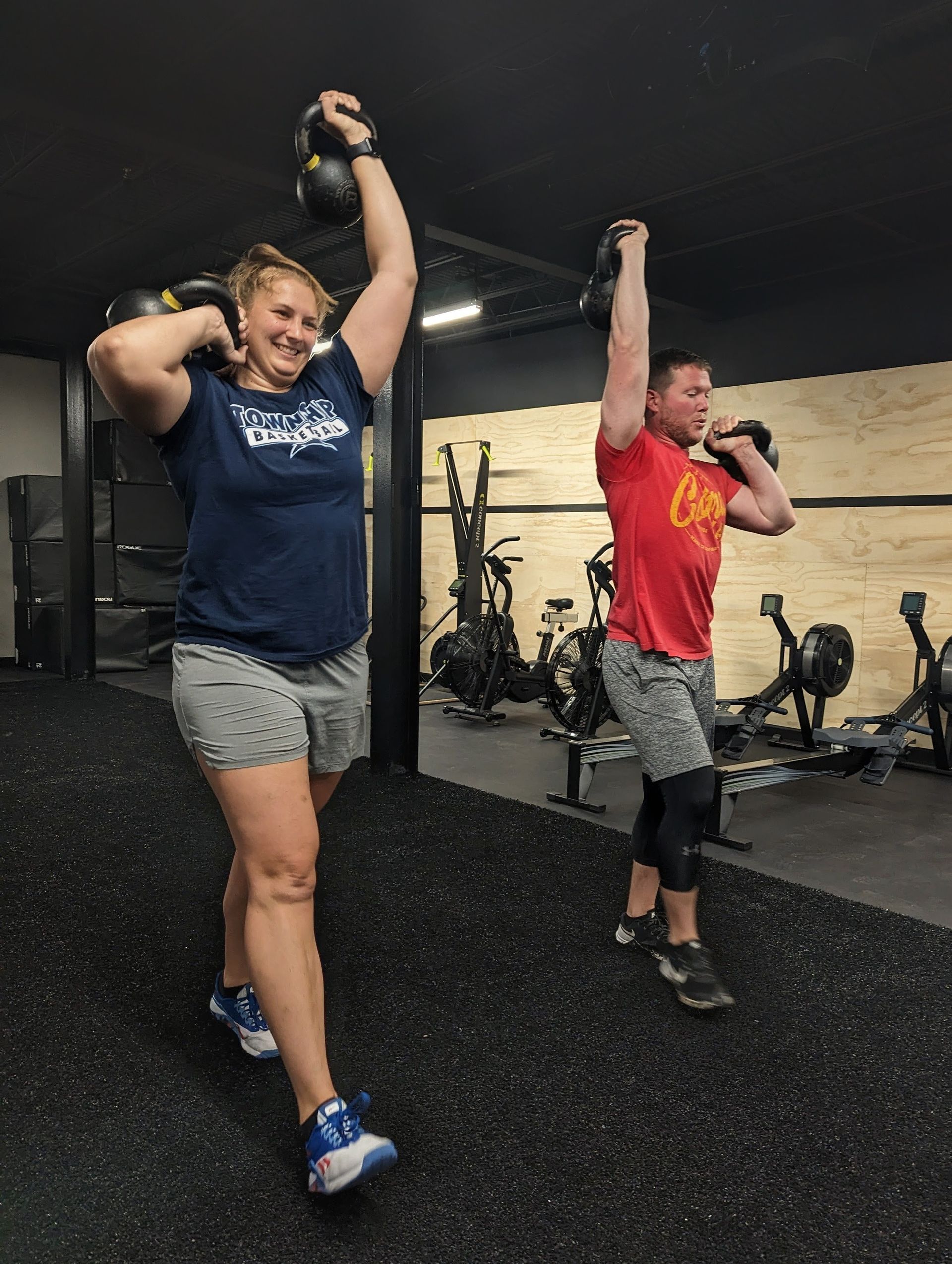Two people exercising in Vitality Athletics in Ephrata, PA, carrying kettlebells overhead. Black floor, neutral background.