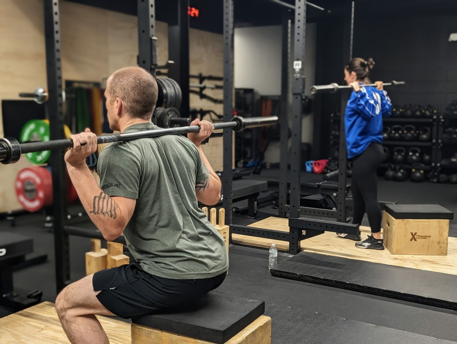 Man in Vitality Athletics in Ephrata, PA squats with barbell on box. Woman squats behind, barbell on back.