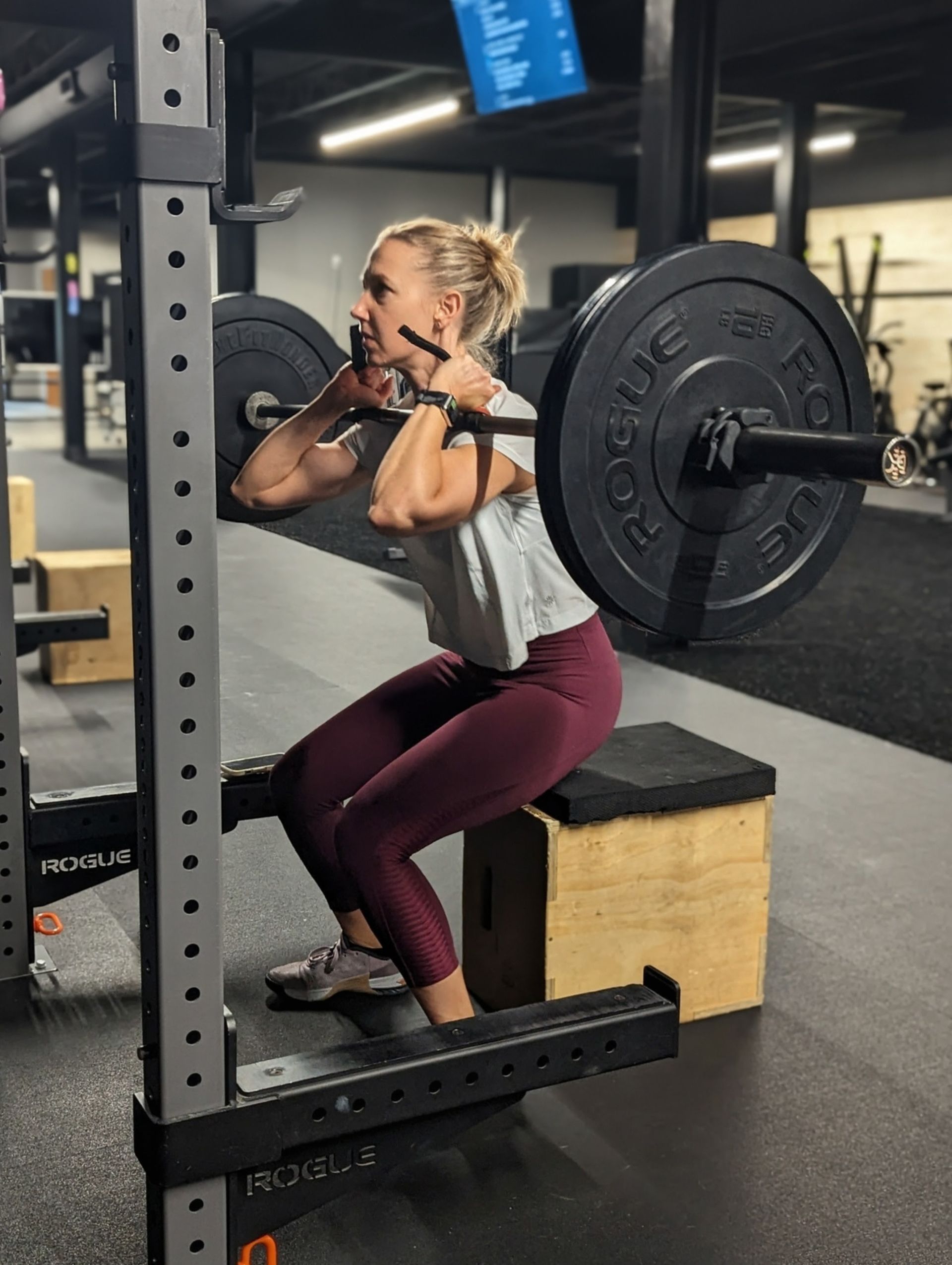 Woman performing a barbell front squat, using a box, at Vitality Athletics in Ephrata, PA. She wears maroon leggings and gray shirt.