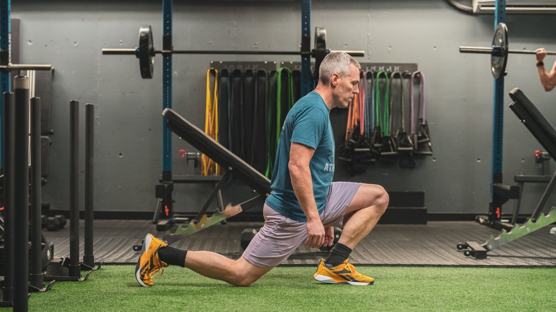 Man in Vitality Athletics in Ephrata, PA, performing a lunge. He has gray hair and is wearing workout attire; gray shorts, a blue shirt, and yellow sneakers.