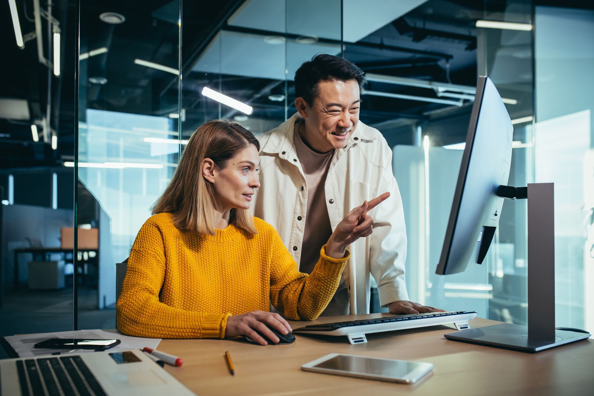 Two coworkers collaborating at a desk in a modern office, looking at a computer screen.