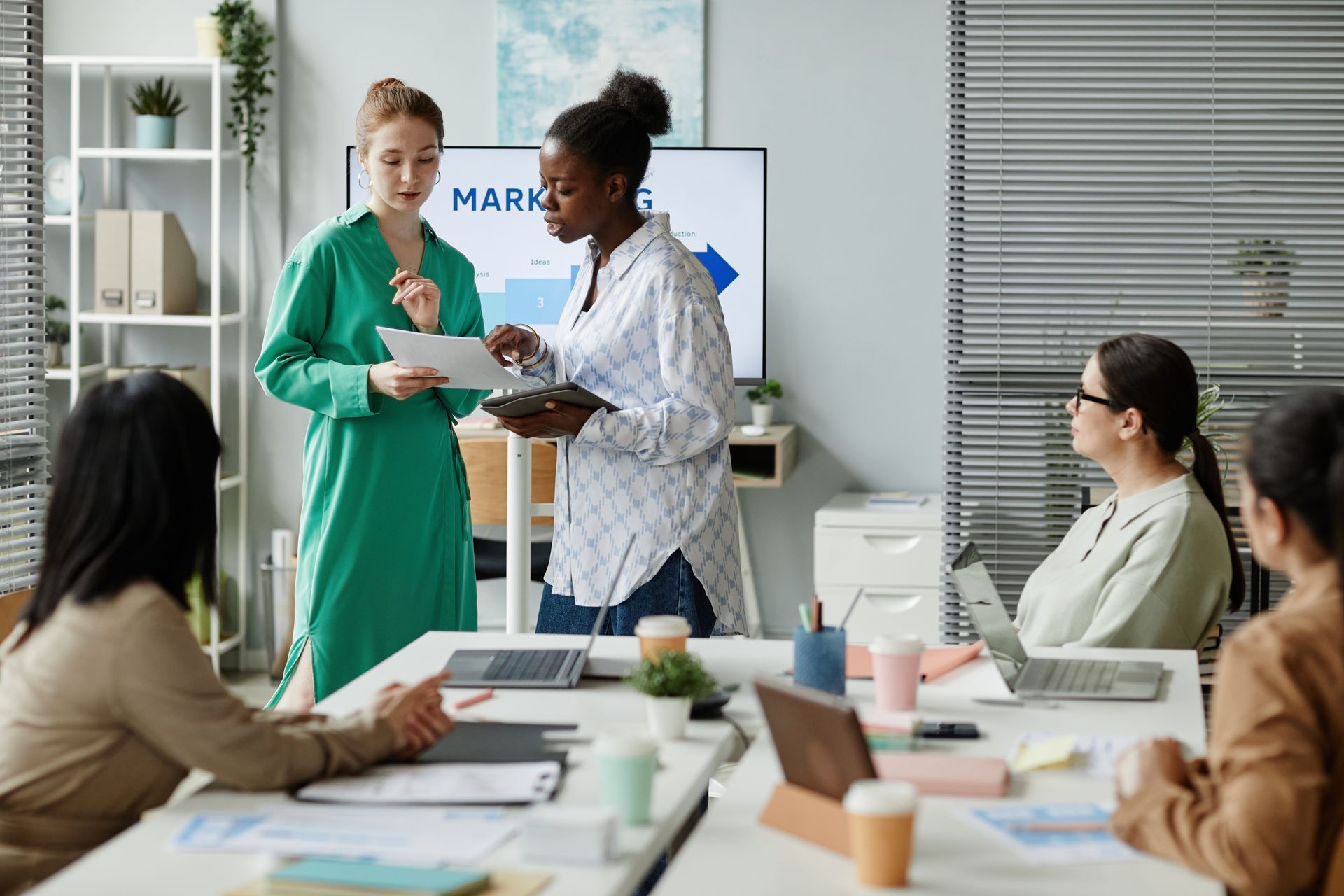 Office meeting with two coworkers reviewing a tablet while others sit around a conference table