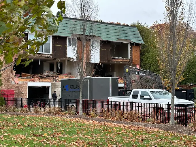 Damaged two-story house with boarded windows and debris in a yard, truck parked in front, autumn trees nearby