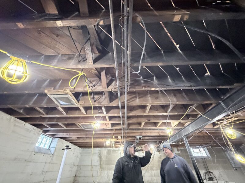 Two workers in hard hats inspect ceiling beams in an unfinished room with exposed wiring.