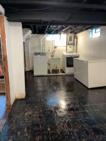 Dim basement room with white appliances, utility fixtures, and dark floor tiles.