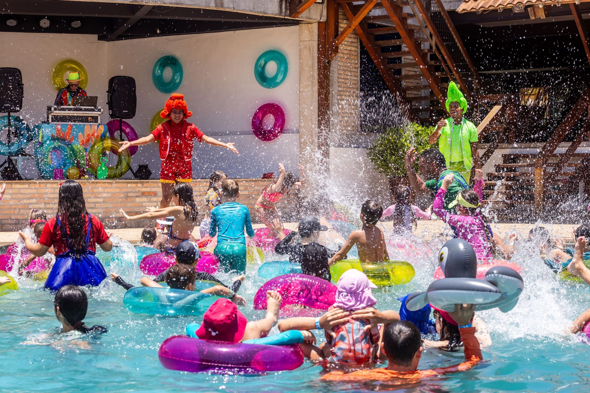 Crianças brincando na piscina com boias coloridas. Os recreadores usam roupas coloridas do lado de fora e animam as crianças. Um dj toca música no palco atrás.