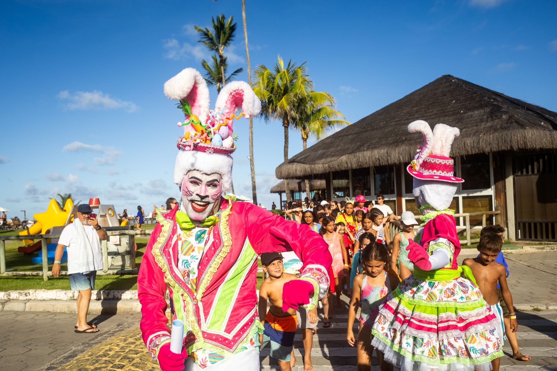 personagens fantasiados de coelho da Páscoa liderando grupo de crianças em atividade ao ar livre no resort, com área externa, coqueiros e hóspedes ao fundo.