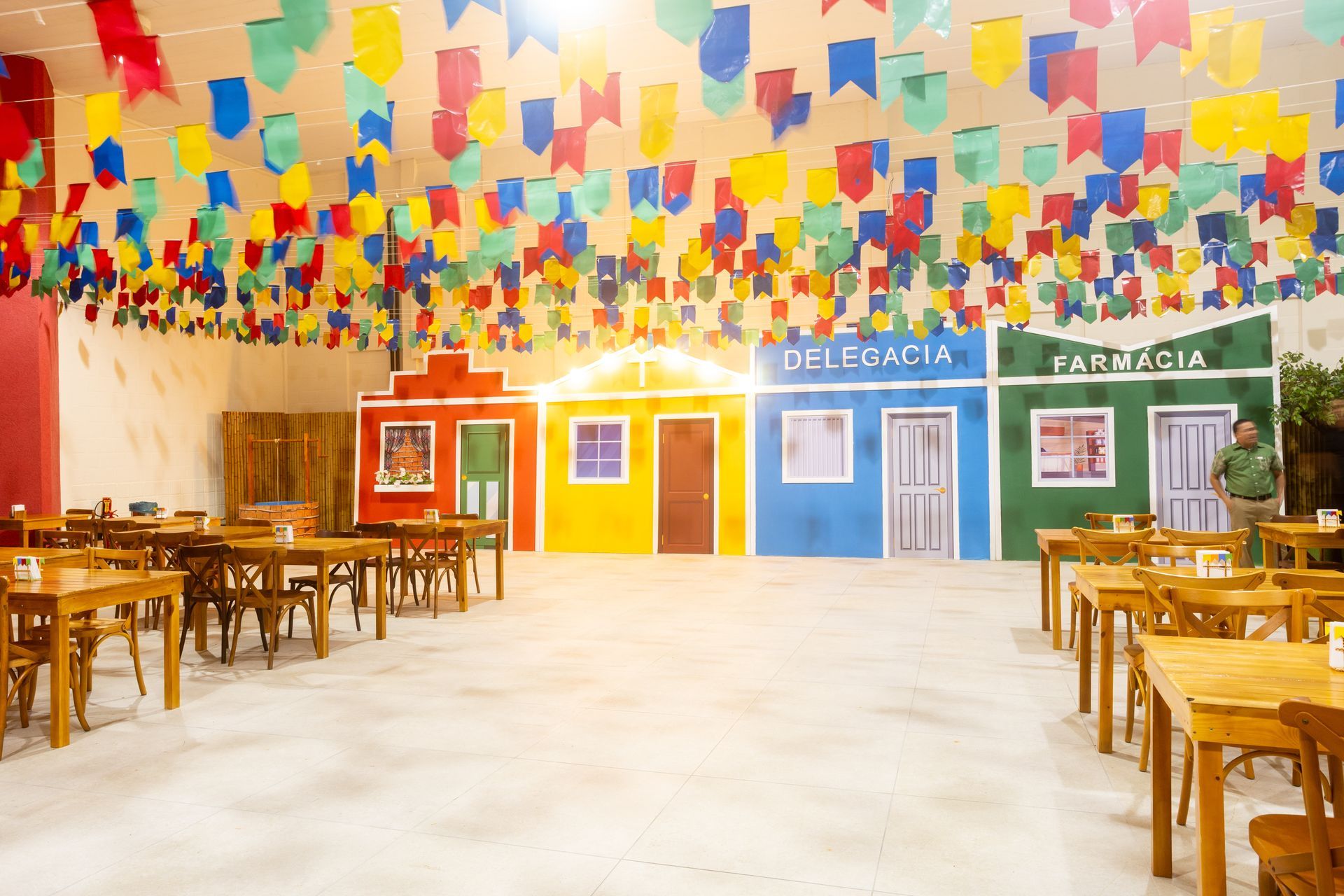 Salão junino com bandeirinhas coloridas, mesas de madeira e fachadas temáticas iluminadas.