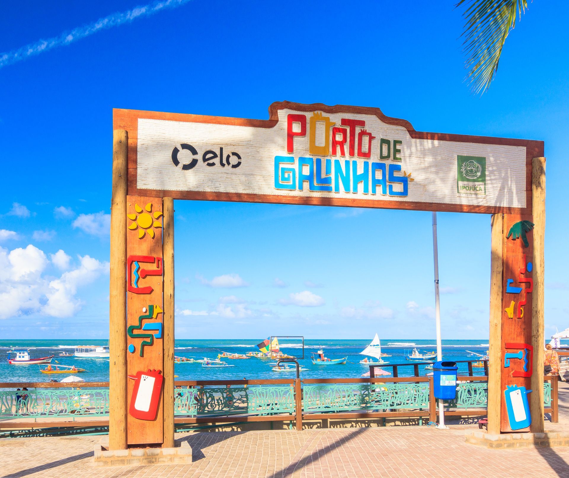 Pórtico de Porto de Galinhas à beira-mar com céu azul, jangadas e cenário tropical em Pernambuco.