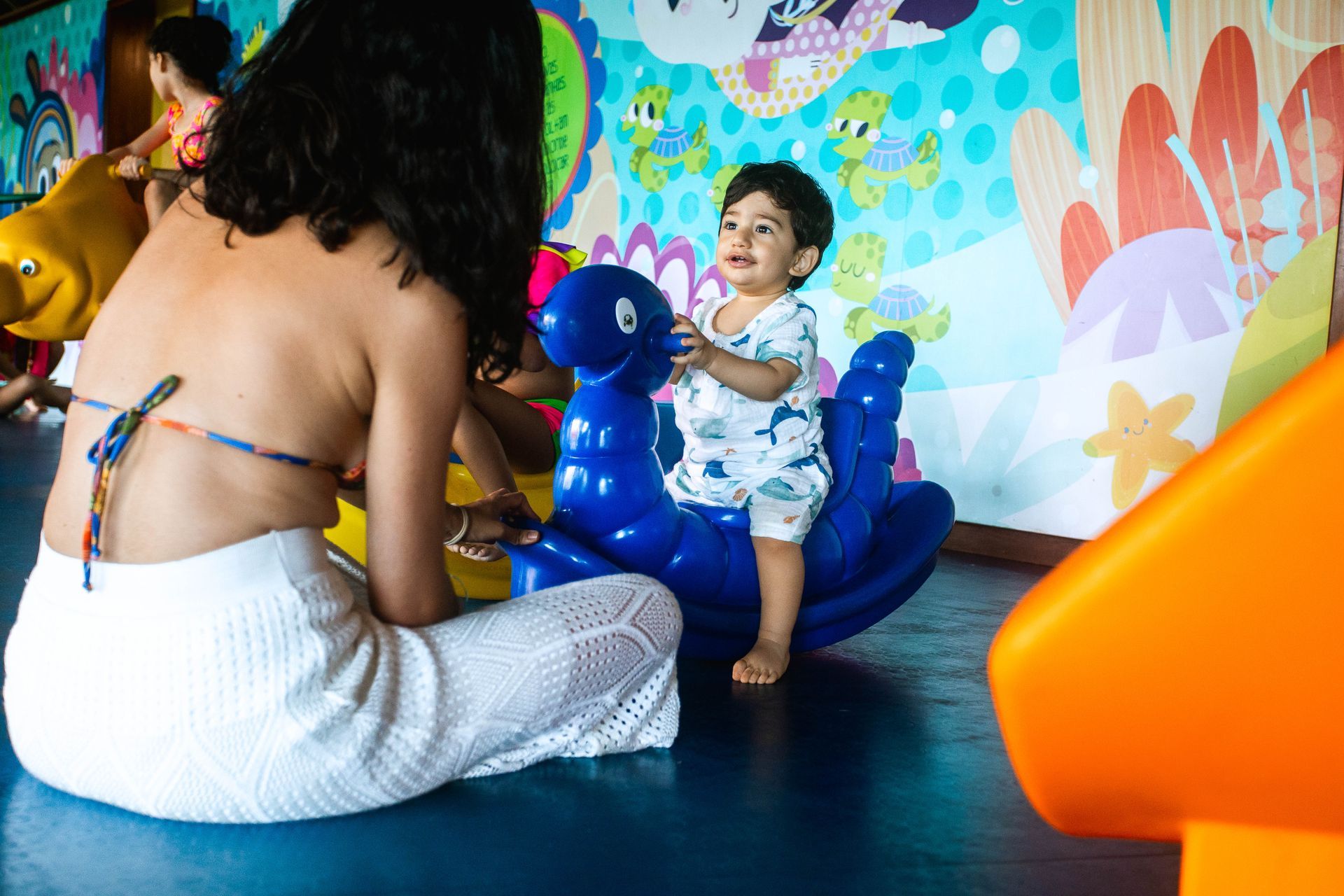 Criança pequena brincando em um cavalinho de balanço azul, acompanhada por um adulto em uma brinquedoteca colorida, com decoração temática marinha e outras crianças ao fundo.