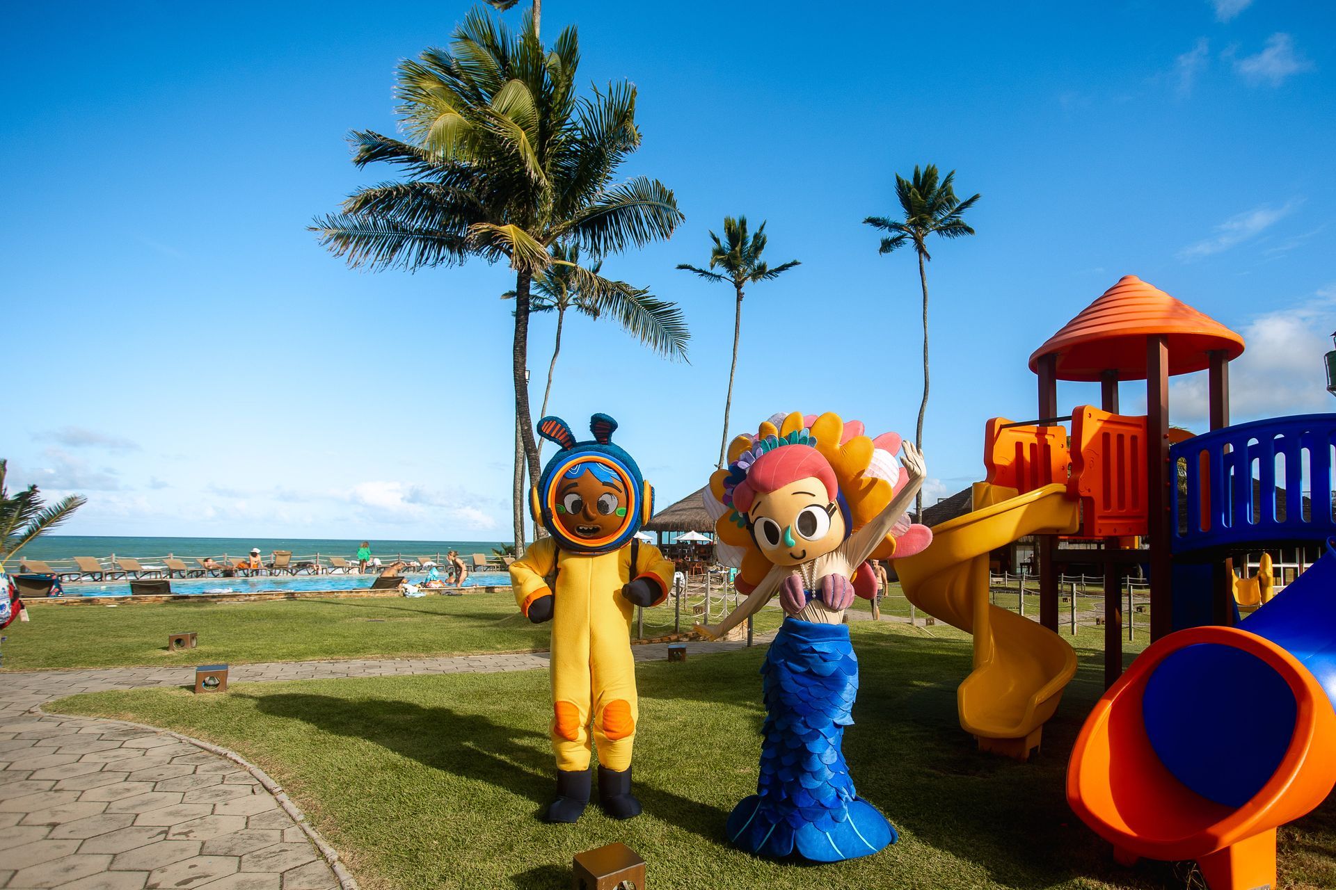 Personagens doArmação Resort no jardim, com piscina, mar e playground ao fundo, em dia de céu azul.