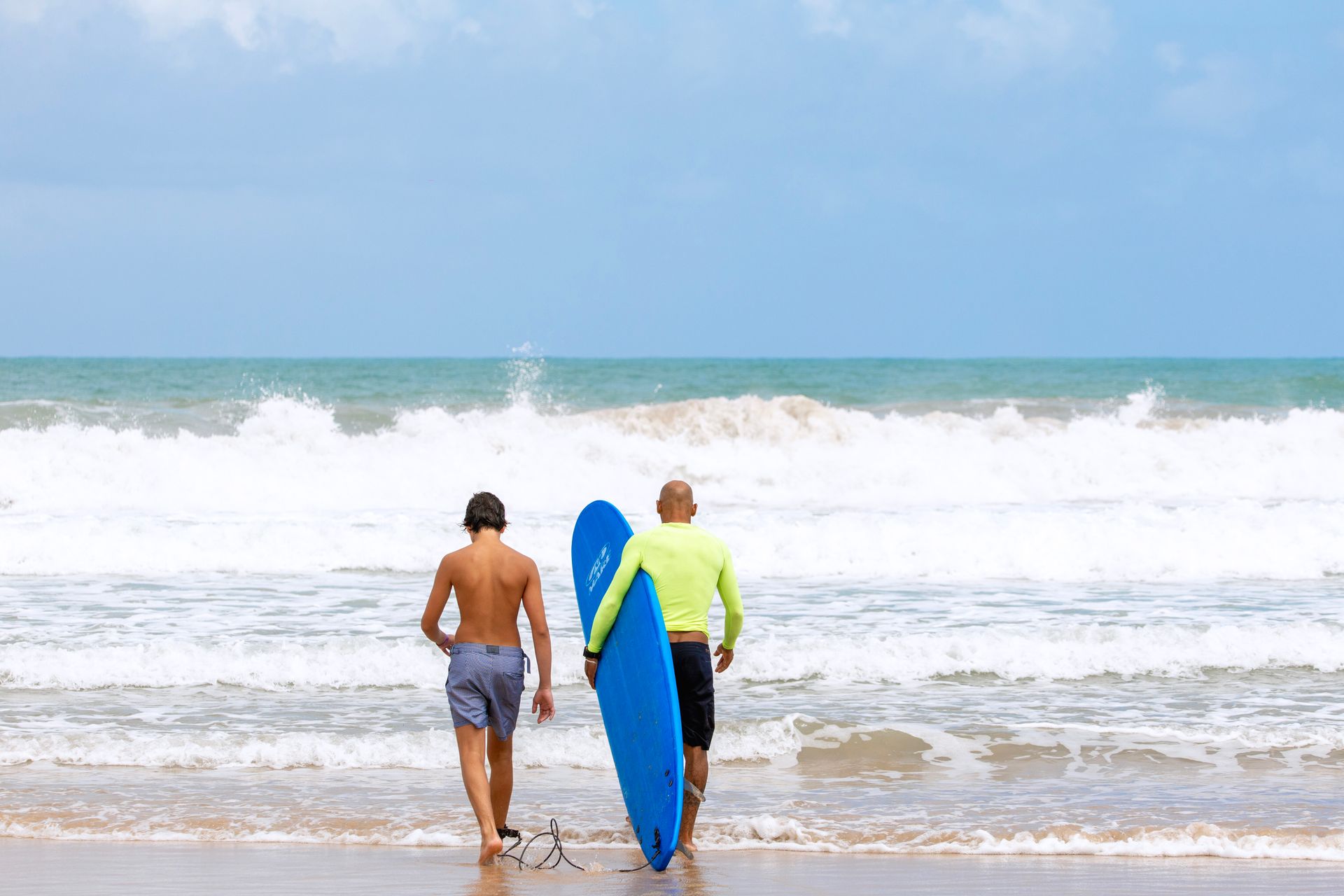 Aula de surf na praia de Porto de Galinhas com instrutor e aluno entrando no mar com uma prancha, mostrando uma das opções de lazer e atividades aquáticas para quem visita a região.