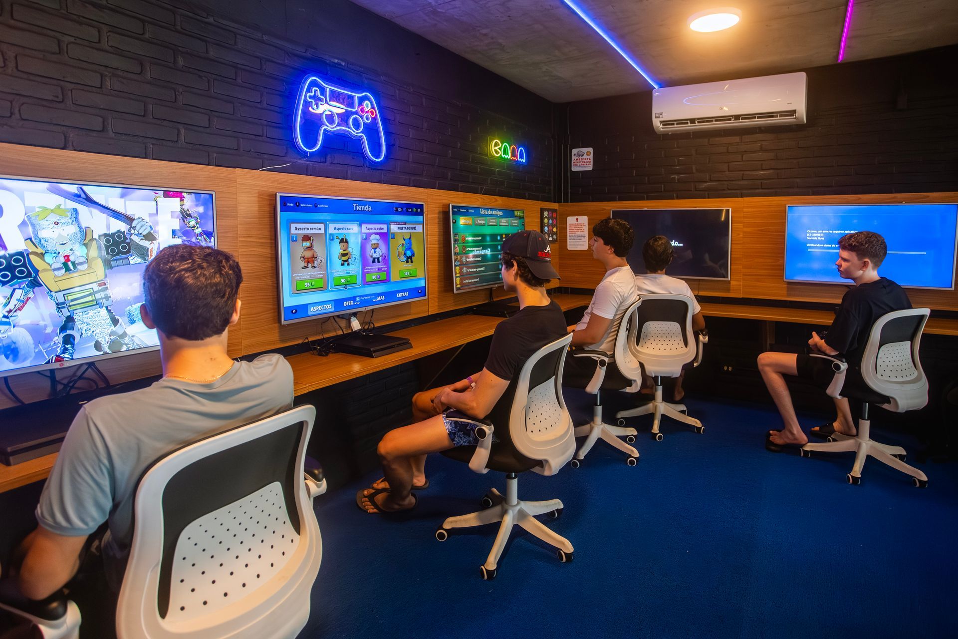 Adolescentes jogando videogame na sala gamer do Armação Resort, com TVs alinhadas e letreiro neon de controle.
