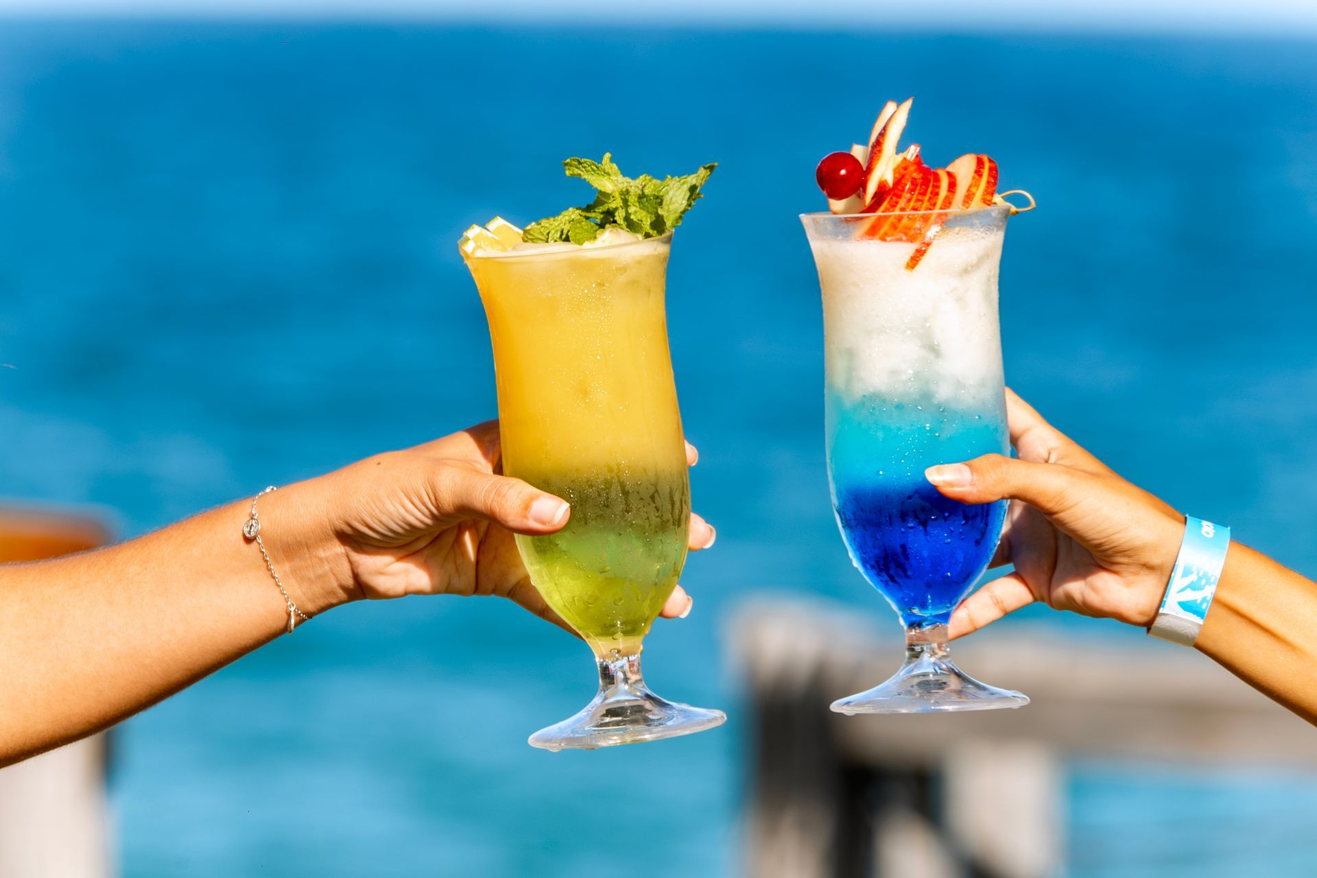 Duas pessoas brindam com drinks coloridos à beira-mar, com o oceano azul ao fundo em um dia ensolarado em Porto de Galinhas.