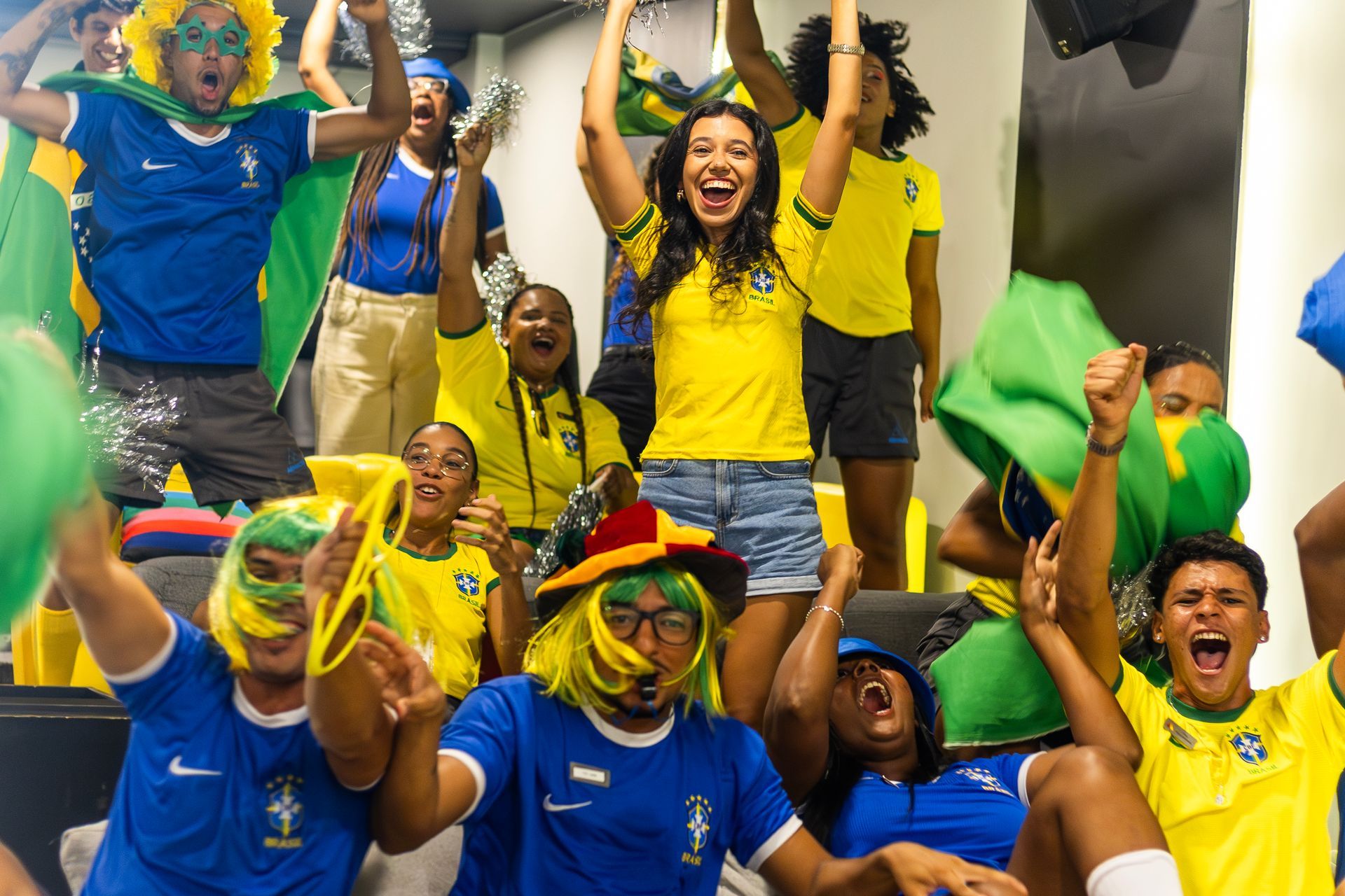Torcedores vestidos com as cores do Brasil comemoram durante a transmissão de uma partida de futebol, em ambiente de entretenimento com clima de torcida e muita animação.
