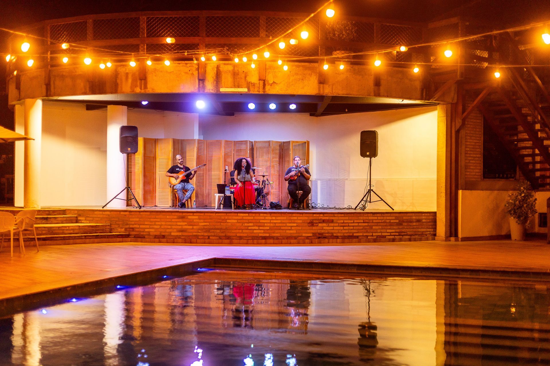Palco do resort com iluminação aconchegante à noite, de frente para uma piscina. Uma cantora e dois músicos no violão e guitarra se apresentam.