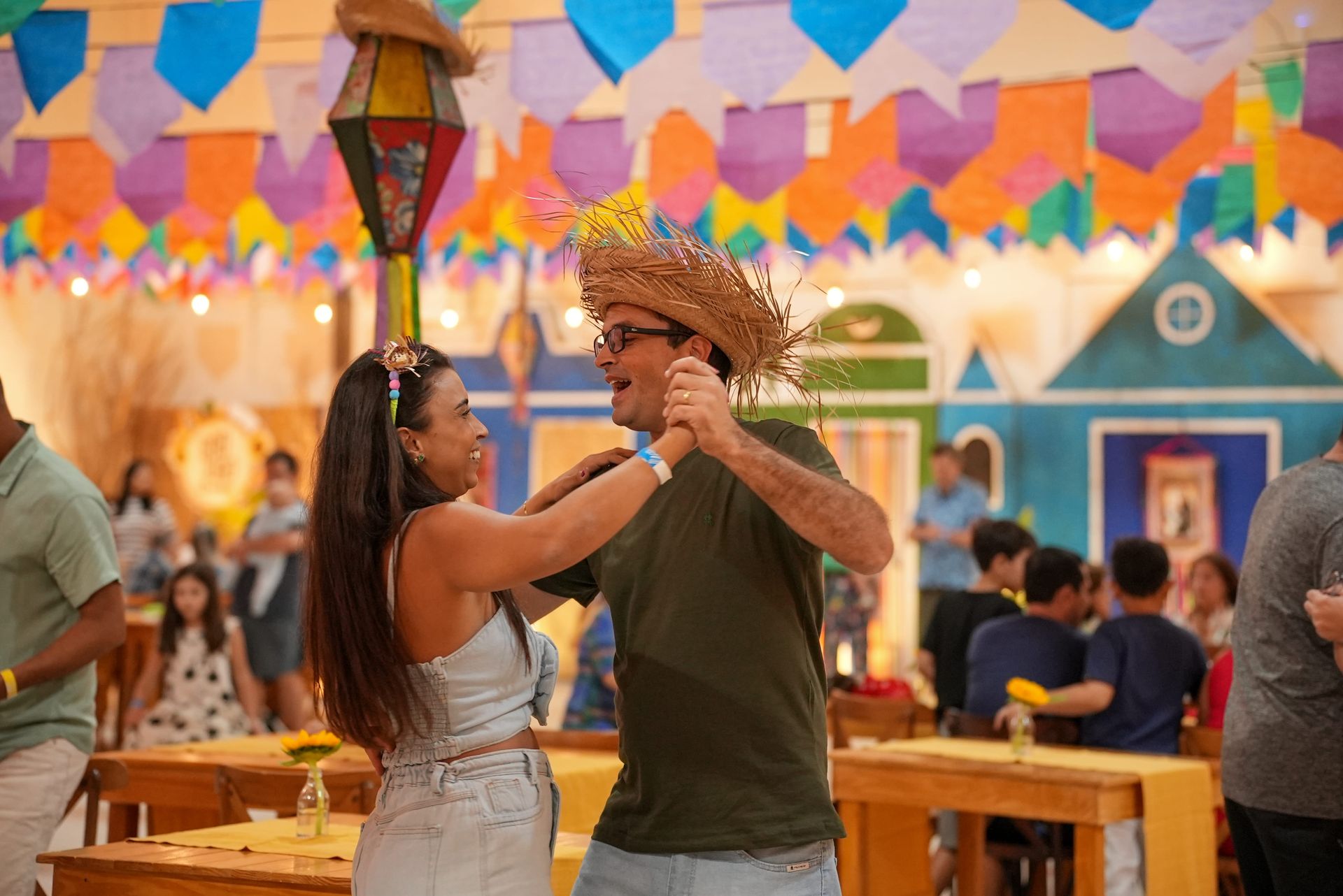 Homem e mulher sorrindo e dançando forró na festa junina, com a cidade cenográfica ao fundo e decoração de bandeiras coloridas no teto. O homem usa um chapéu de palha.