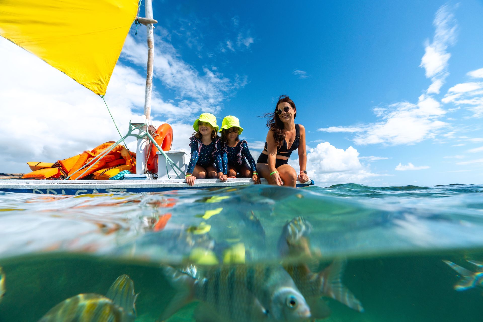 Mãe e duas filhas pequenas, gêmeas, estão sentadas em jangada no mar verde e cristalino. Na água é possível ver peixes. O céu está azul e o dia ensolarado.