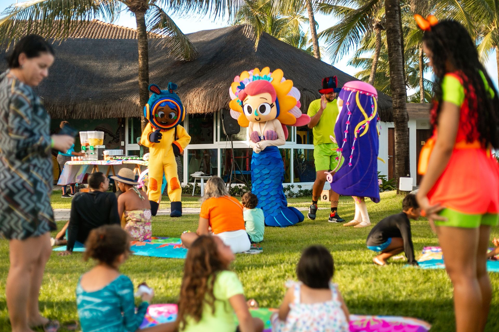 Crianças fazem piquenique no gramado do Armação Resort. Elas estão sentadas em toalhas coloridas e assistem os personagens Lui, Pérola e Cora dançando em pé.