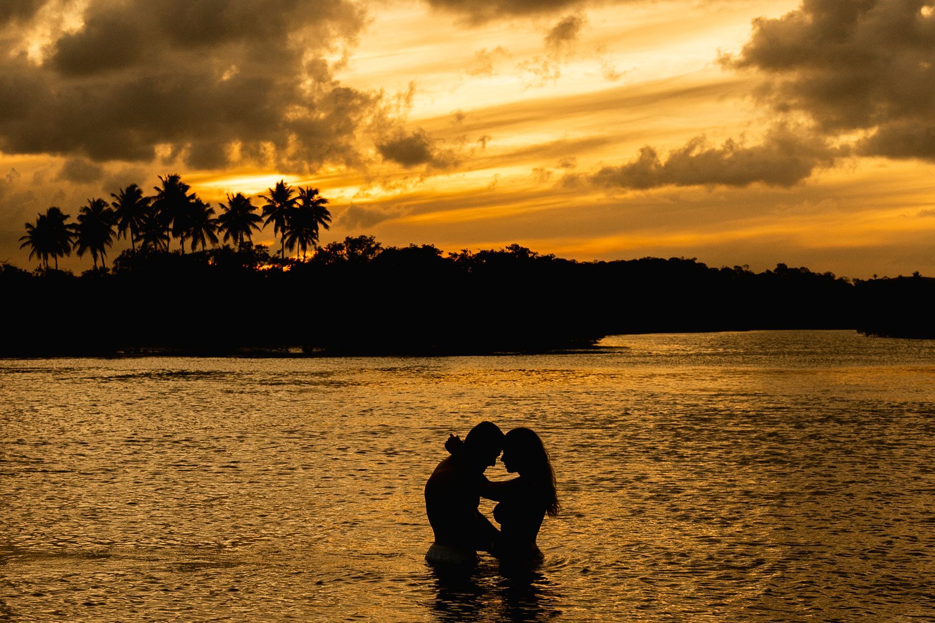 Silhueta de casal na água durante pôr do sol dourado no pontal de maracaípe com coqueiros ao fundo.