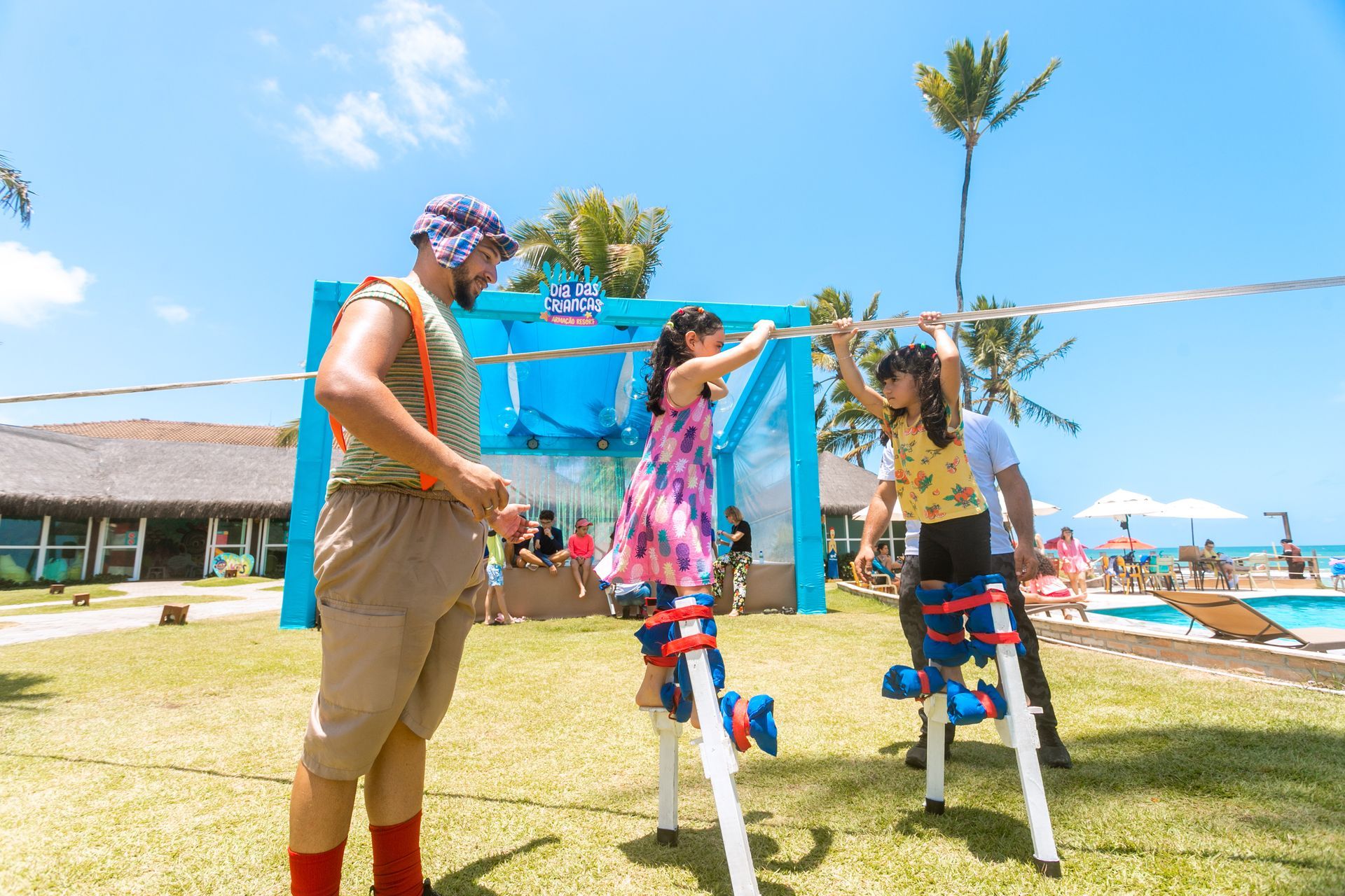 Brincadeira com tio e duas meninas em perna de pau, em oficina de circo no gramado do resort. Ao ar livre, o dia está ensolarado e as crianças se divertem.