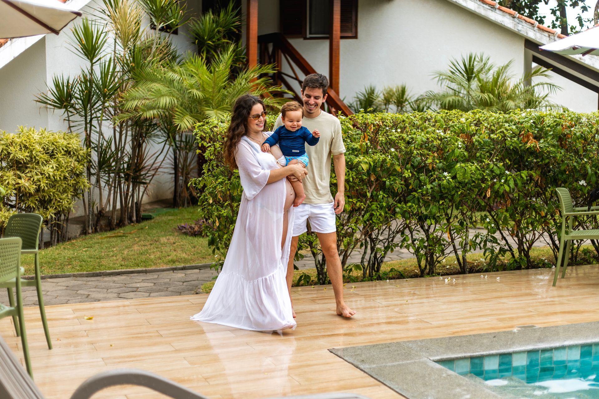 Família feliz com mãe grávida e bebê caminhando à beira da piscina do resort.
