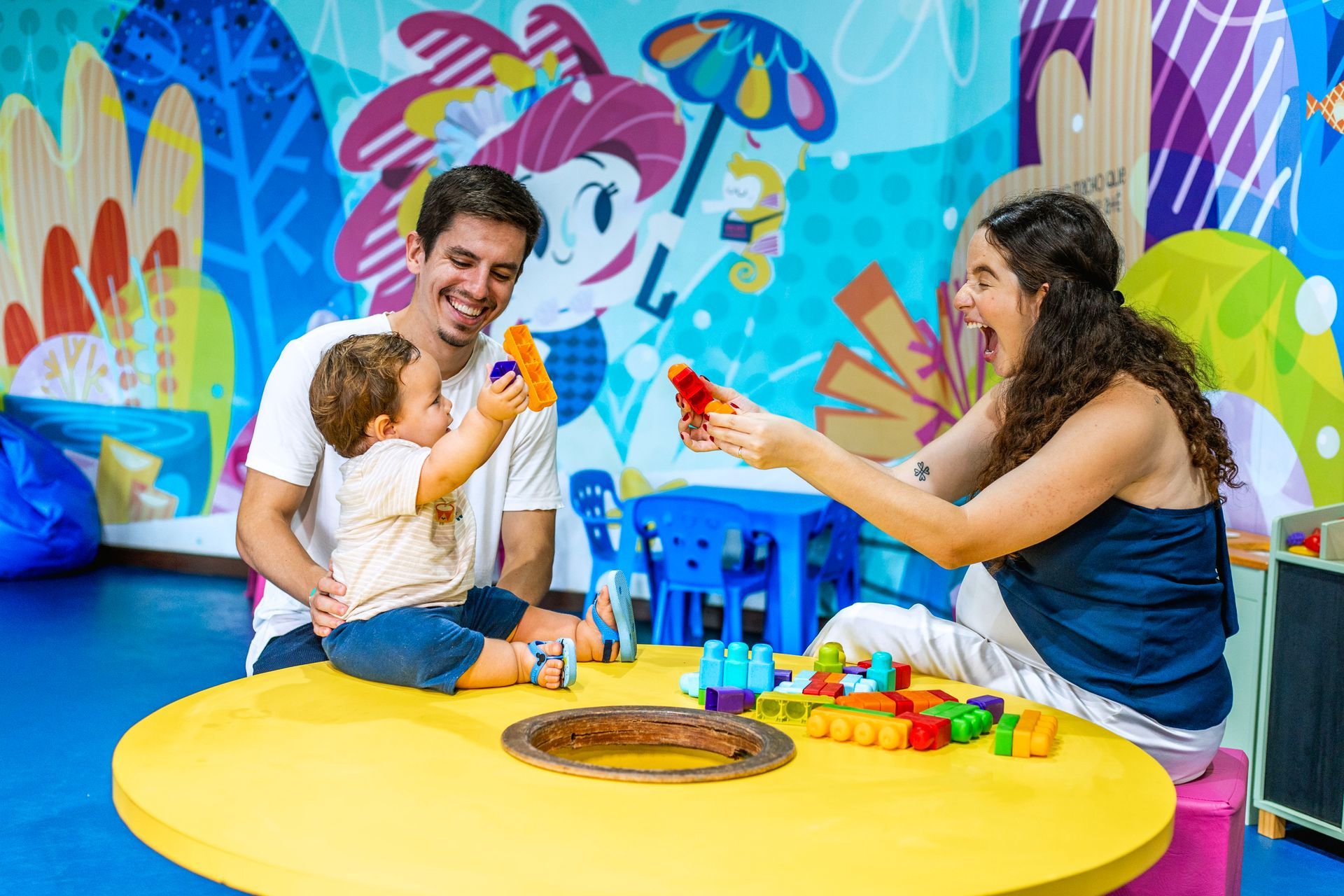 Família sorridente brincando com bebê e blocos no espaço kids do resort.