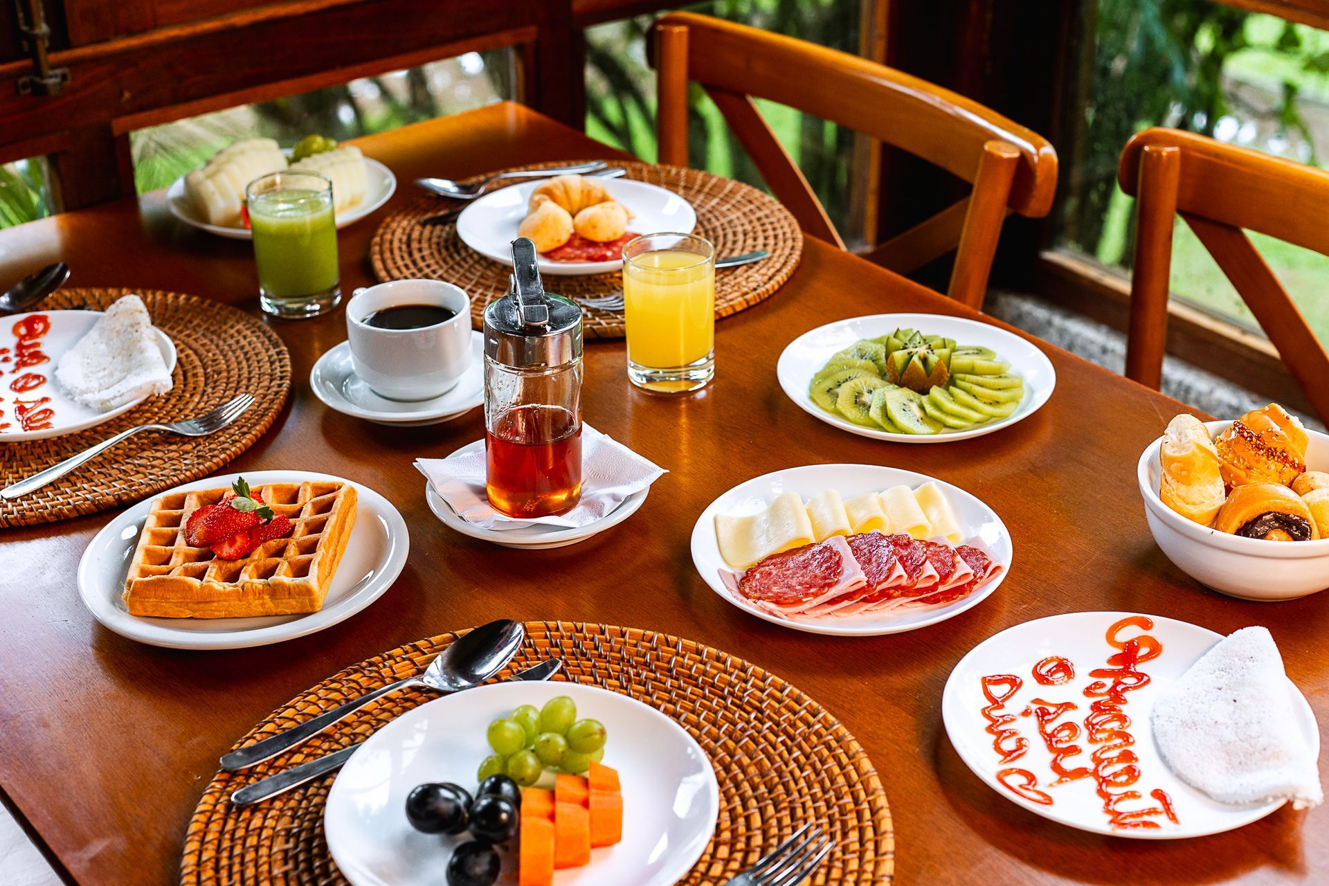 Café da manhã com frutas, pães, queijos, frios, waffles, sucos e café servido à mesa.