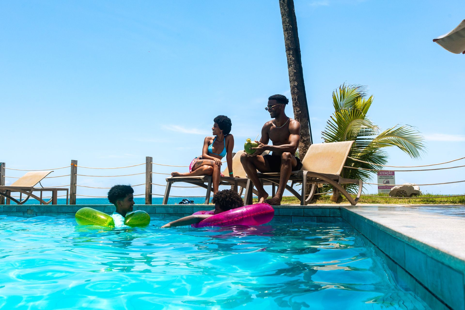 Família aproveitando a piscina do Armação Resort em Porto de Galinhas, com adultos relaxando nas espreguiçadeiras e crianças brincando na água em um dia ensolarado.