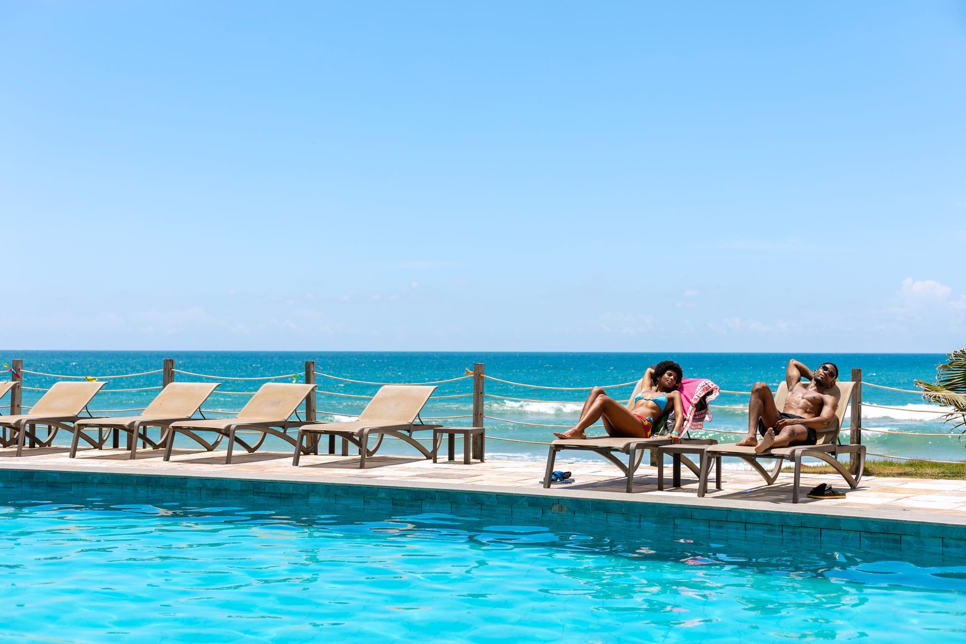 Piscina à beira-mar com espreguiçadeiras e hóspedes aproveitando um dia ensolarado.