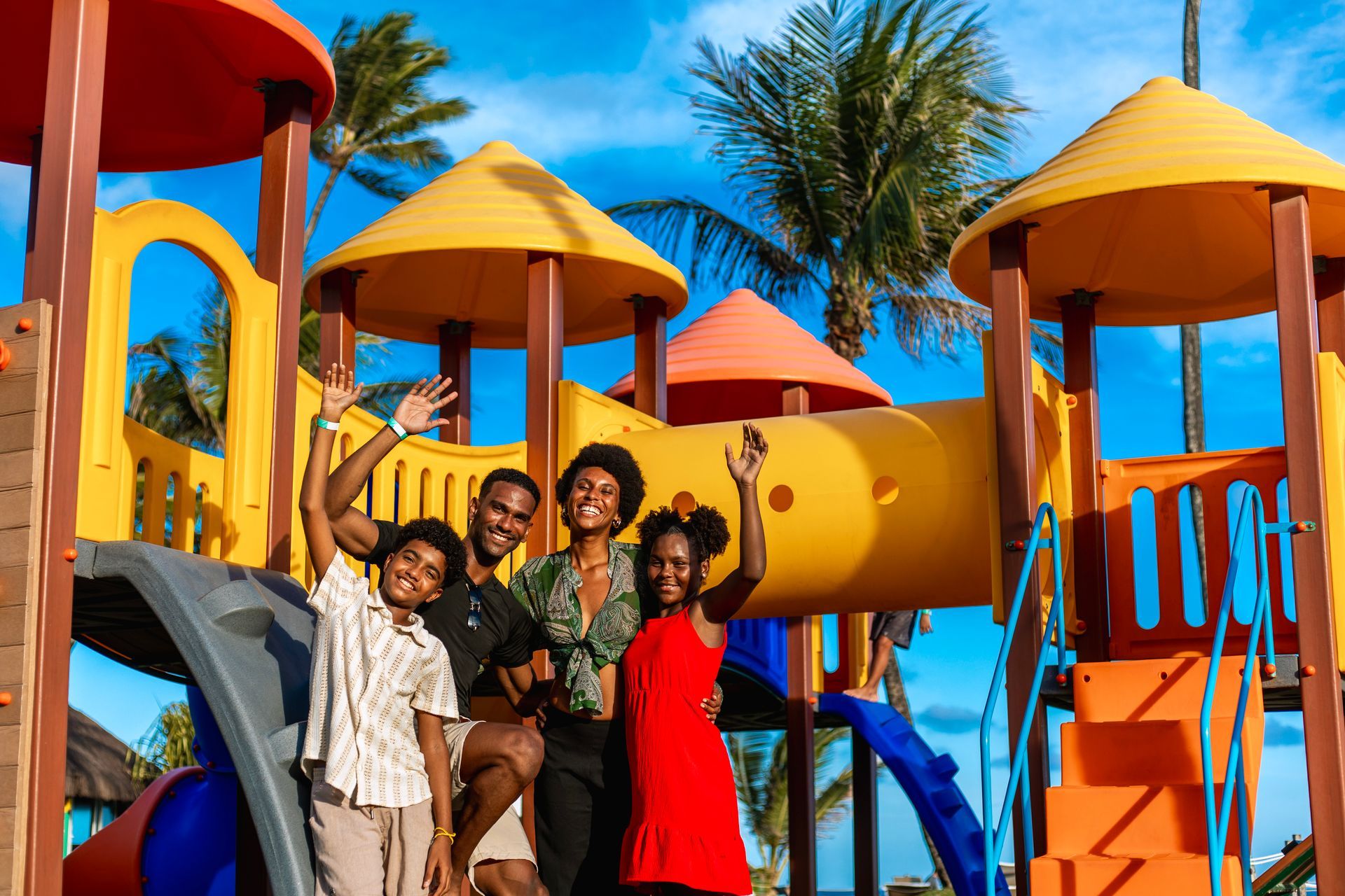 Família feliz posando em playground infantil colorido com coqueiros ao fundo.