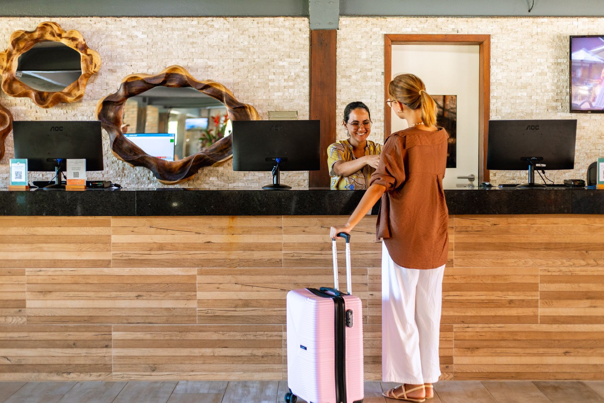 Recepcionista atende hóspede com mala no balcão da recepção do Armação Resort.