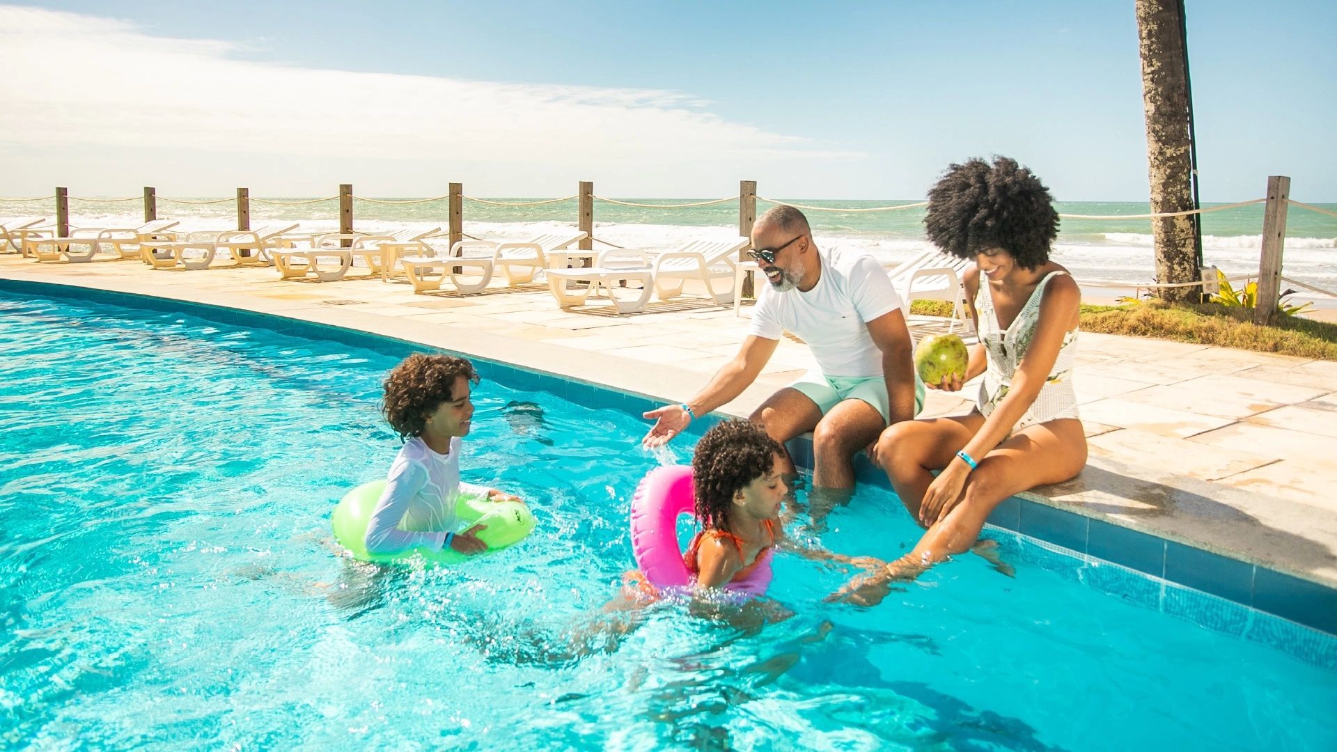 Família se divertindo à beira da piscina, com as crianças brincando na água e os pais sentados relaxando, em um resort à beira-mar.