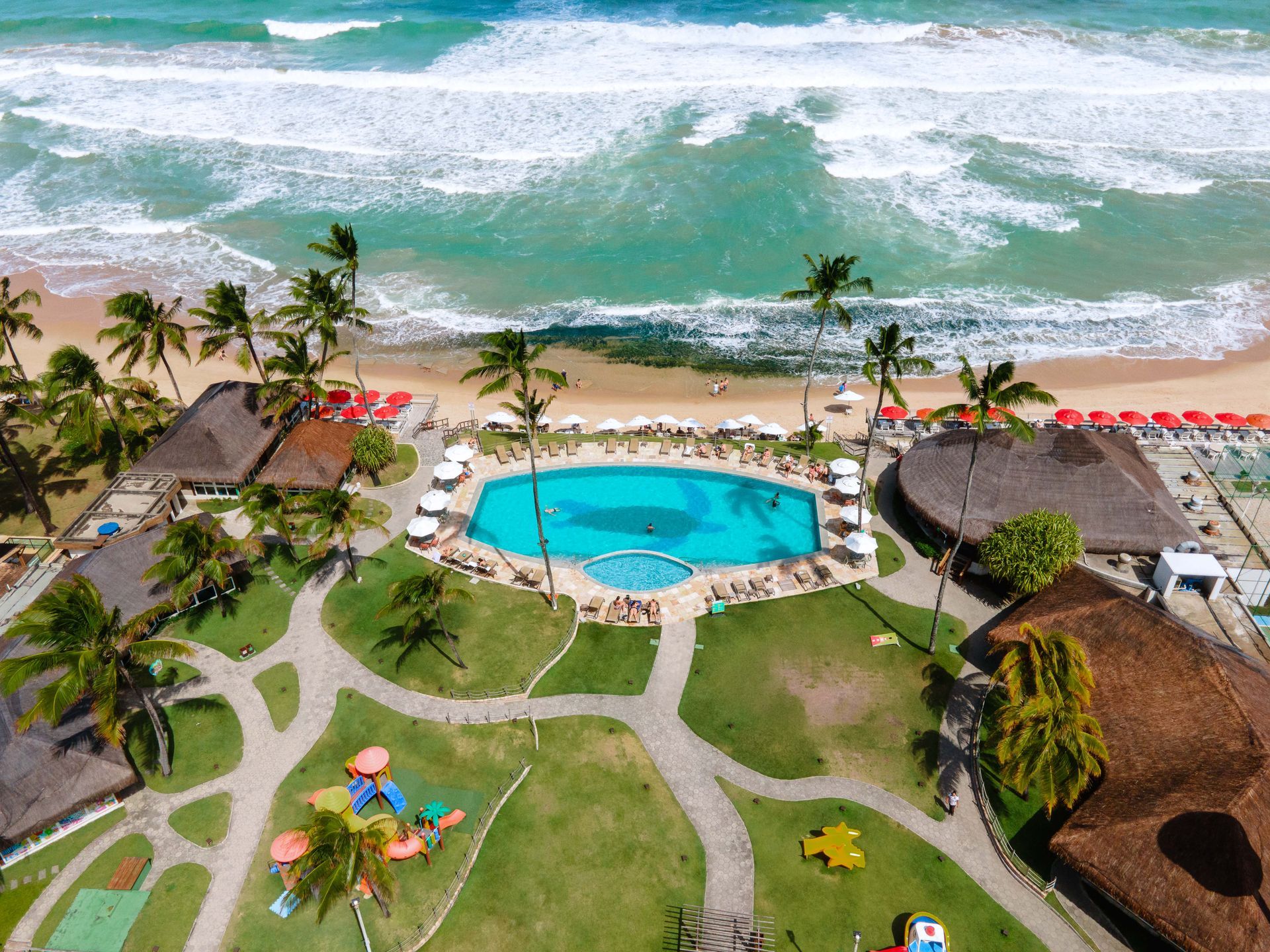 Vista aérea do Armação Resort mostra a piscina azul cercada de áreas verdes, estruturas de palha e praia com mar cristalino.