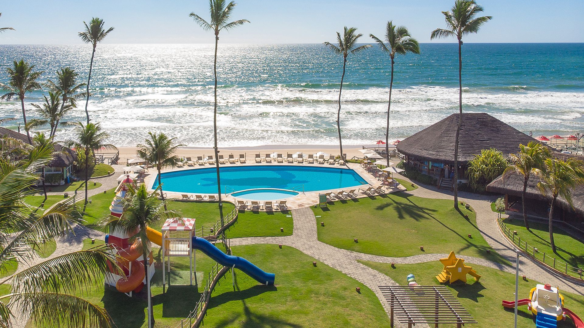 Vista aérea da piscina do Armação Resort à beira-mar, com tobogã infantil e cadeiras de sol alinhadas ao redor, destacando o mar de Porto de Galinhas ao fundo.